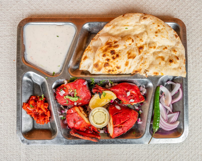 Indian thali with tandoori chicken, naan, raita, and pickled vegetables on a metal tray.