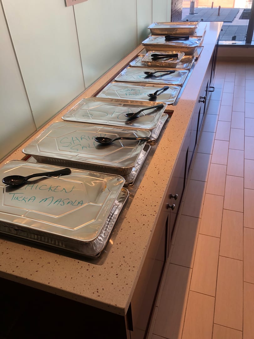 A buffet line of foil-covered catering trays with handwritten labels like "Chicken Tikka Masala" and "Saag Paneer".
