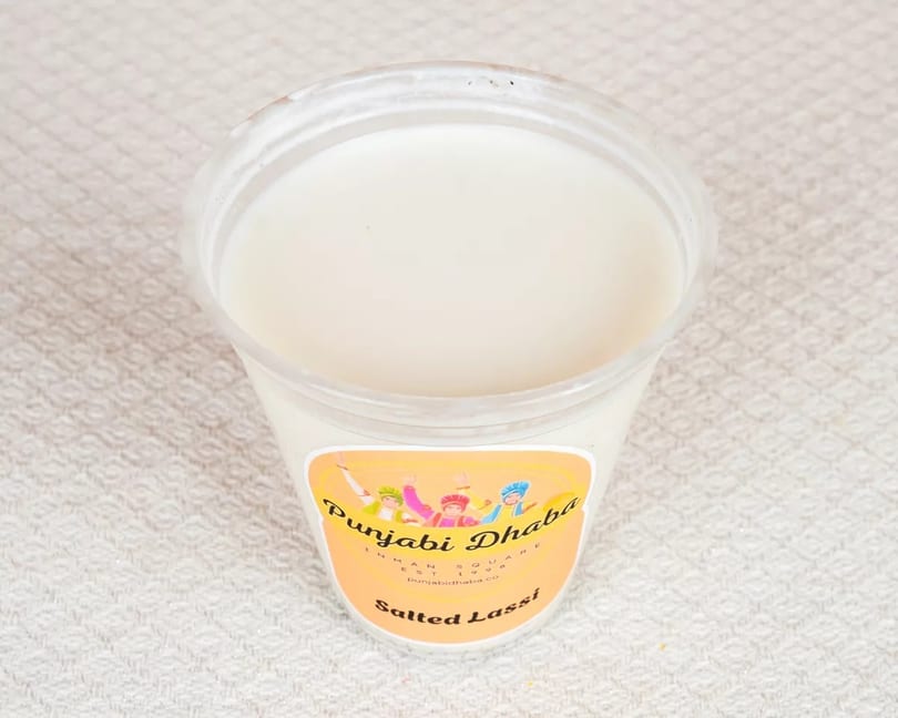 A clear plastic cup of white salted lassi from Punjabi Dhaba on a textured white surface.