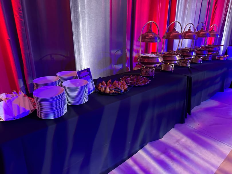 A buffet table with chafing dishes, plates, and appetizers under red and blue lighting.