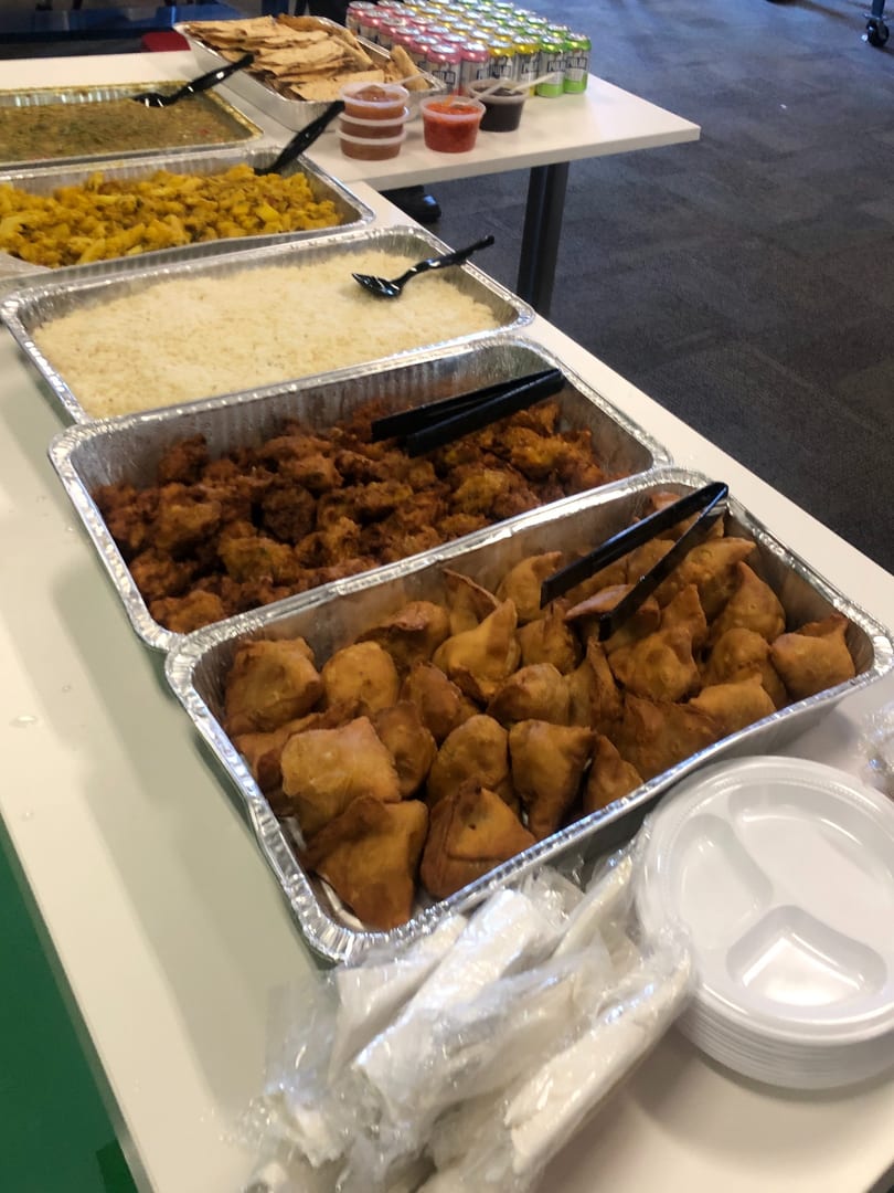 A buffet line with trays of Indian food, including samosas, rice, and curries, with drinks and plates.